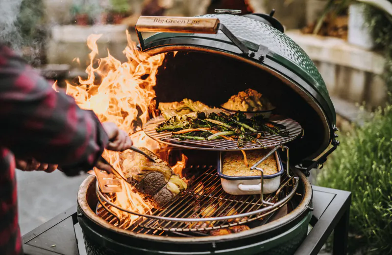 Person grilling meat and vegetables on an open Big Green Egg ceramic grill with flames