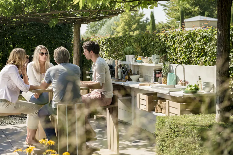 Four adults chatting at a modern outdoor kitchen bar under a leafy pergola
