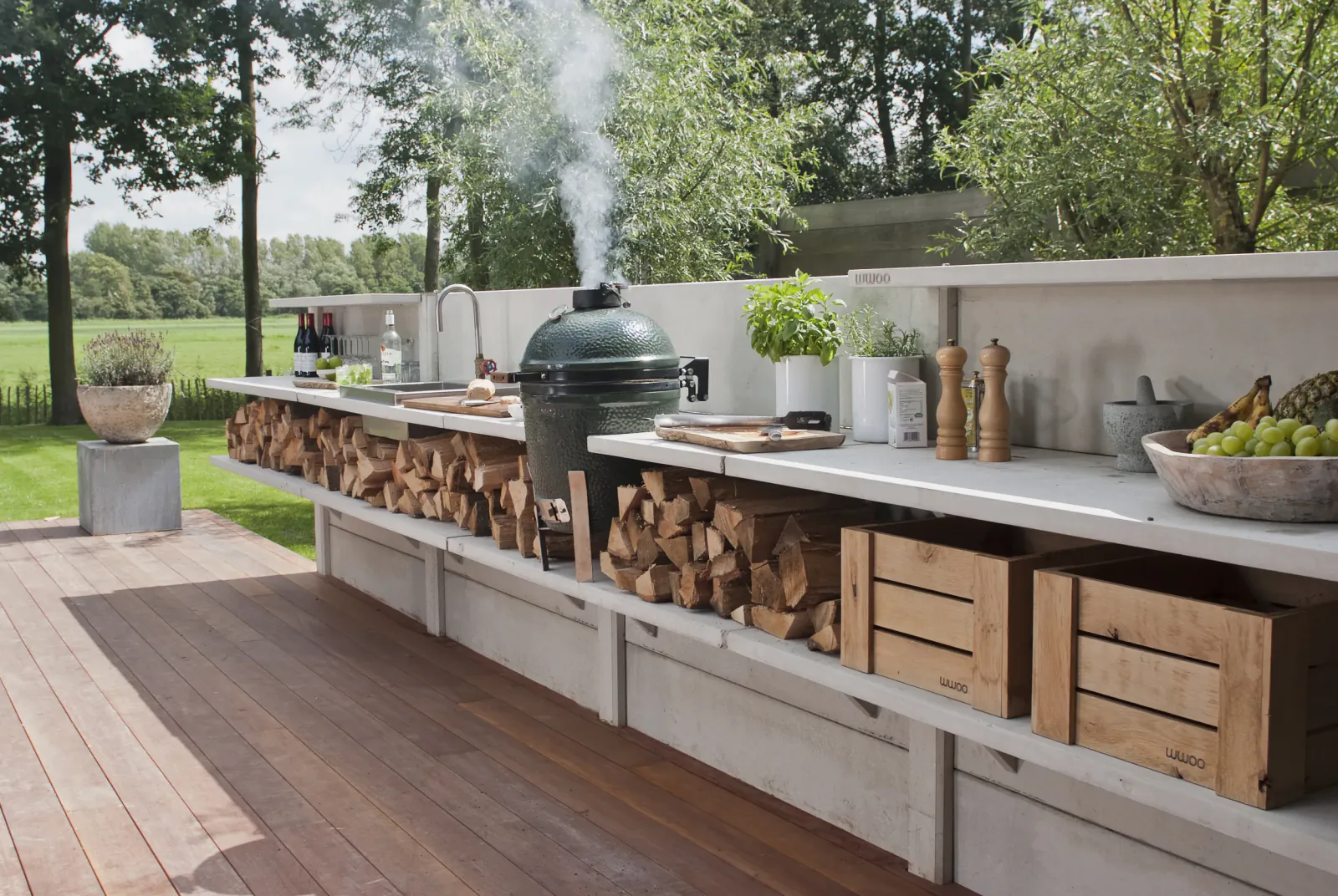 Modern outdoor kitchen on a wooden deck with a smoking green grill, stacked firewood, sink, and fresh produce in a garden setting