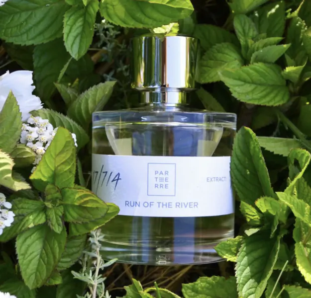 Clear glass perfume bottle labeled 'RUN OF THE RIVER' by PAR TERRE EXTRACTS, nestled among green leaves and white flowers