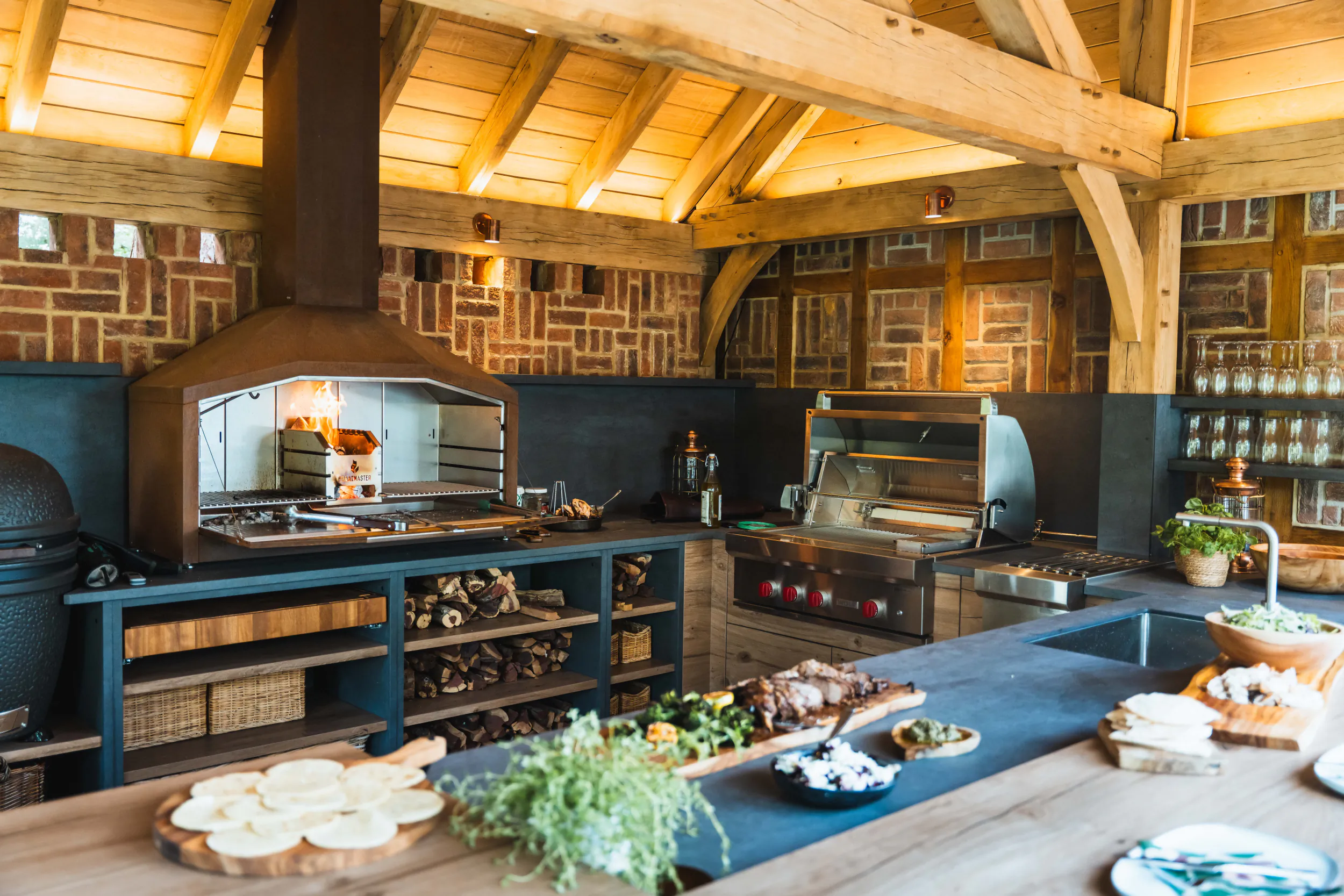 Rustic wooden outdoor kitchen with brick walls, wood-fired oven, grill, and fresh food prep on a large counter