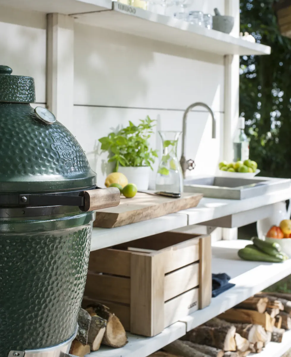Outdoor kitchen with green ceramic grill, wooden counter, sink, fresh herbs and vegetables