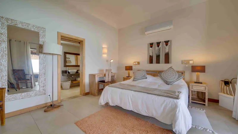 Bright modern bedroom with white bed, patterned pillows, wooden furniture, large ornate mirror, and doorway to bathroom