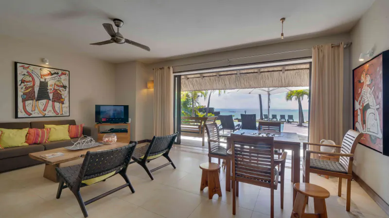 Modern open-plan living room with ocean-view balcony, wooden dining table, grey sofa with colorful cushions, and two large African-style wall artworks