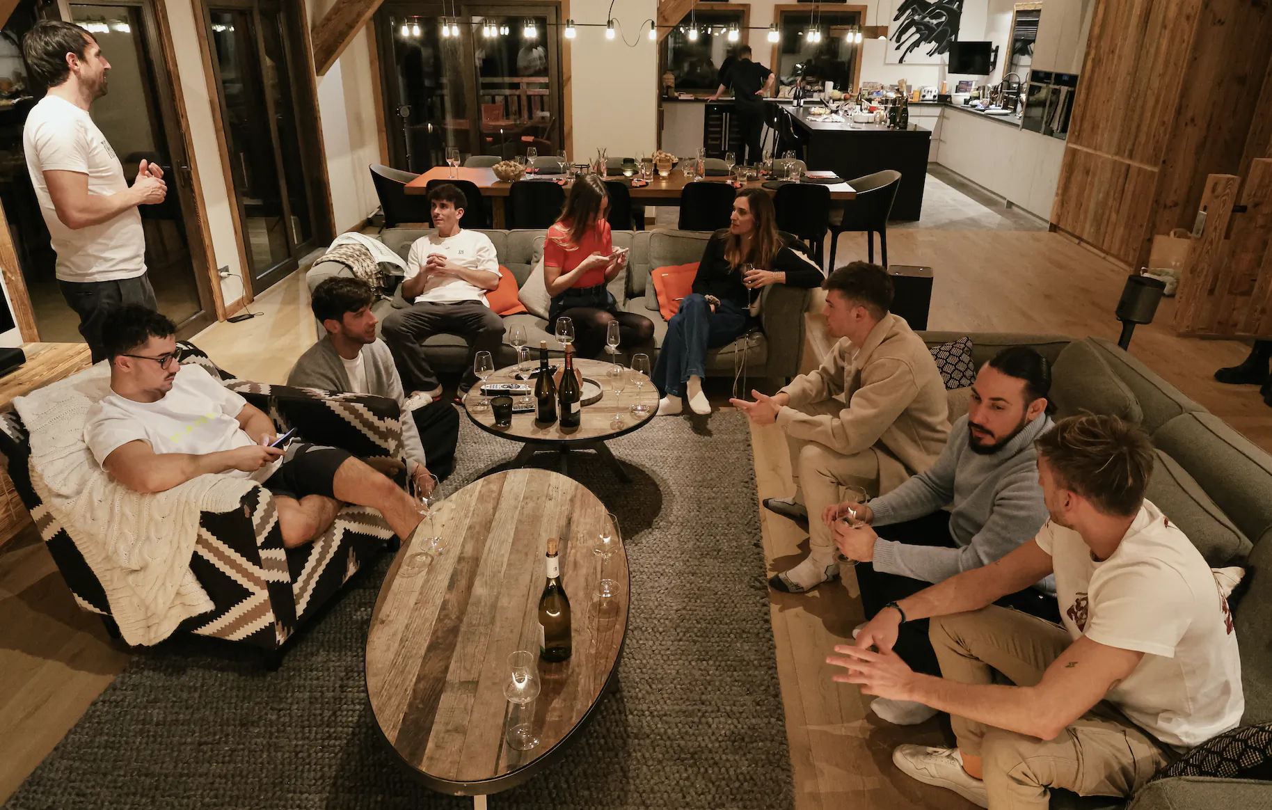 Friends gathered in a modern open-plan living room, chatting and drinking wine around wooden coffee tables