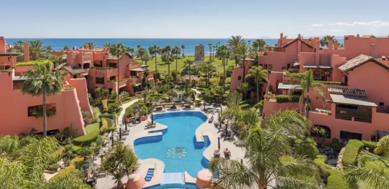 Aerial view of a luxury resort with pink buildings, large blue pool, palm trees, and ocean backdrop in Marbella