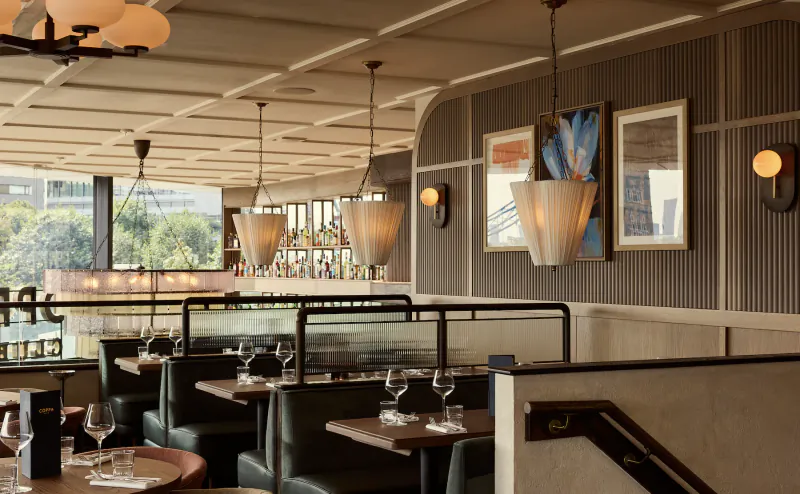 Modern restaurant interior with dark leather booths, wooden tables set with wine glasses, pendant lights, and large windows overlooking trees