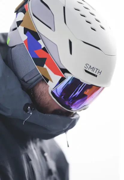 Close-up of a person wearing a white Smith ski helmet with colorful geometric design and purple-tinted goggles, looking down in snowy conditions