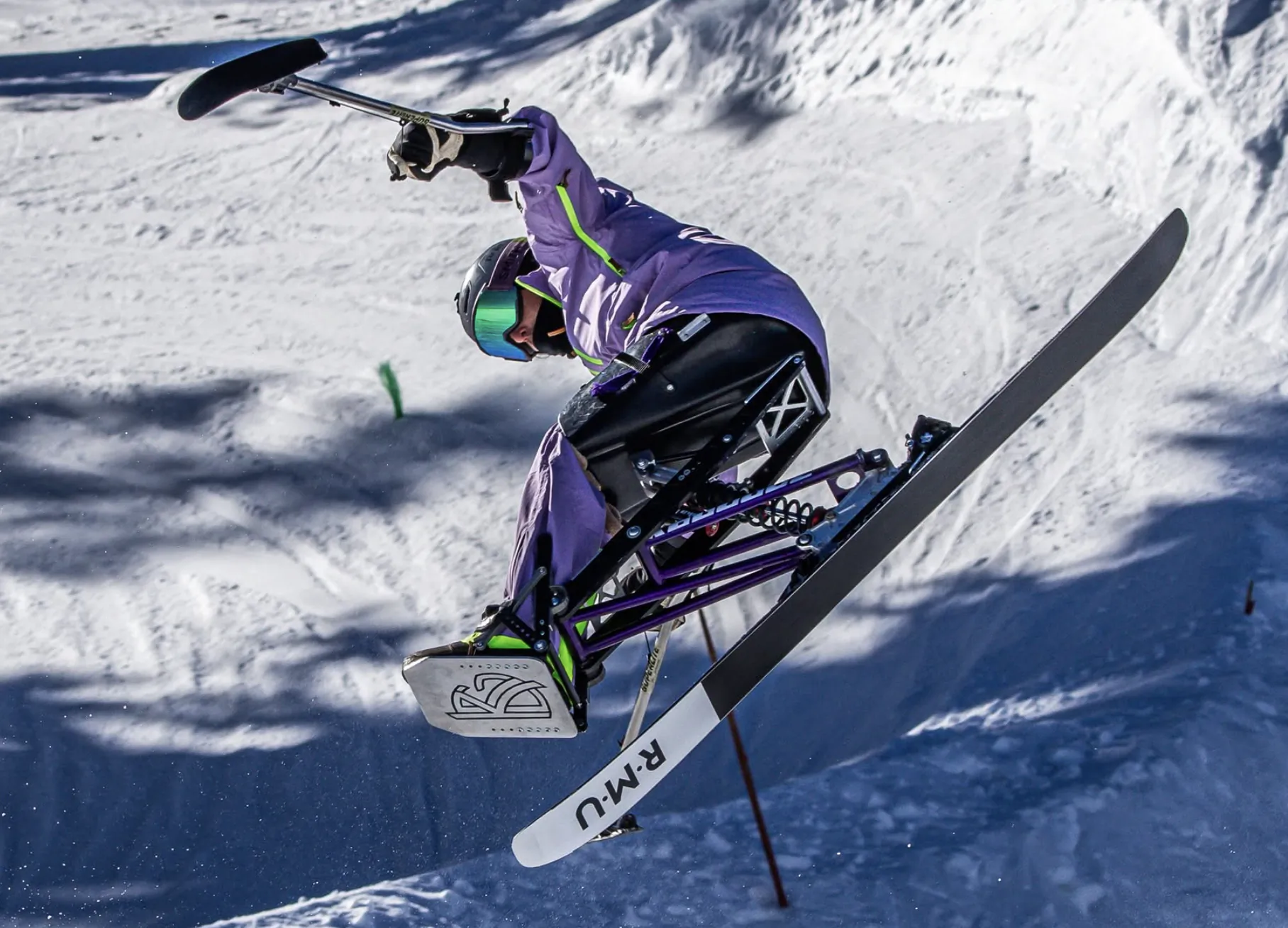Adaptive skier in purple jacket performing aerial trick on sit-ski with RMU branding in snowy mountains