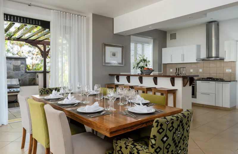 Modern open-plan dining area with a long wooden table set for eight, green and patterned chairs, overlooking a kitchen and outdoor patio