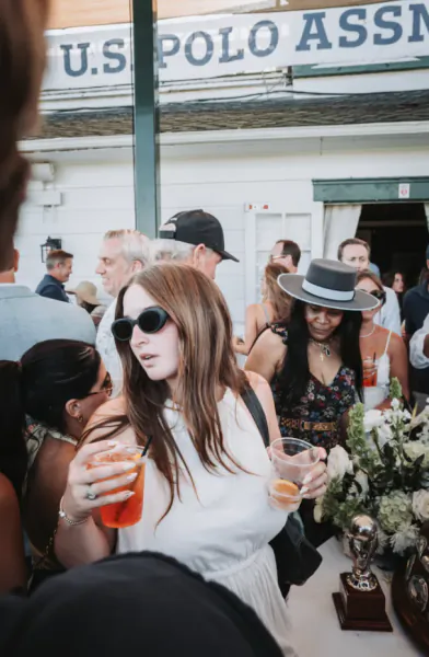 Woman in white dress holding cocktails at U.S. Polo Assn. event with crowd and trophy table
