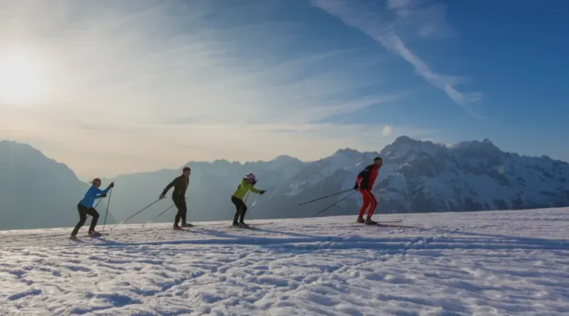 Four skiers in colorful jackets skiing across a vast snowy mountain slope under a bright blue sky