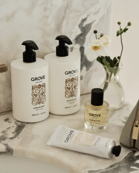 Grove England Anemone skincare products: two white pump bottles, small glass perfume, white tube, and white flower in vase on marble surface
