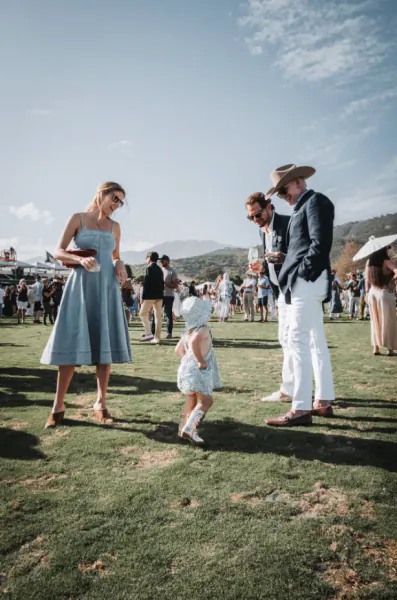 Woman in blue dress and man in white pants with hat watch toddler in sun hat on grass at outdoor polo event