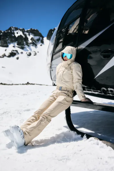 Woman in cream ski suit and blue goggles sits on black helicopter skid in snowy mountain landscape