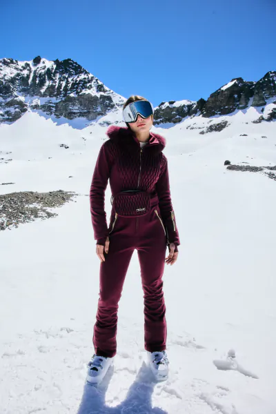Woman in maroon ski suit and goggles standing on snowy mountain slope under blue sky