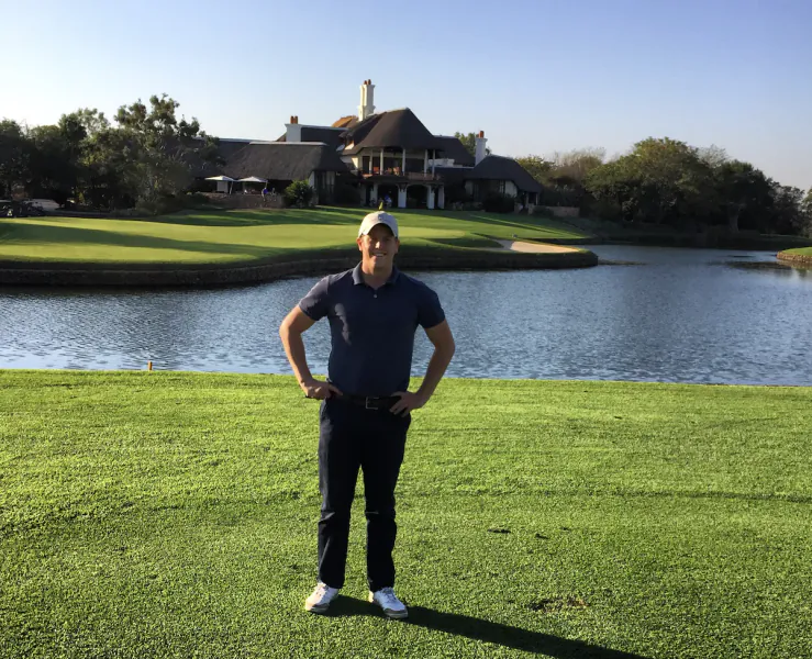 Man in navy polo and cap stands with hands on hips on a golf course green, with a lake and large thatched-roof clubhouse behind him