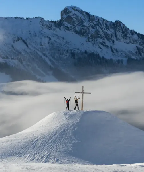 Two hikers celebrate beside a wooden summit cross on a snowy peak above clouds, with jagged mountains in the background