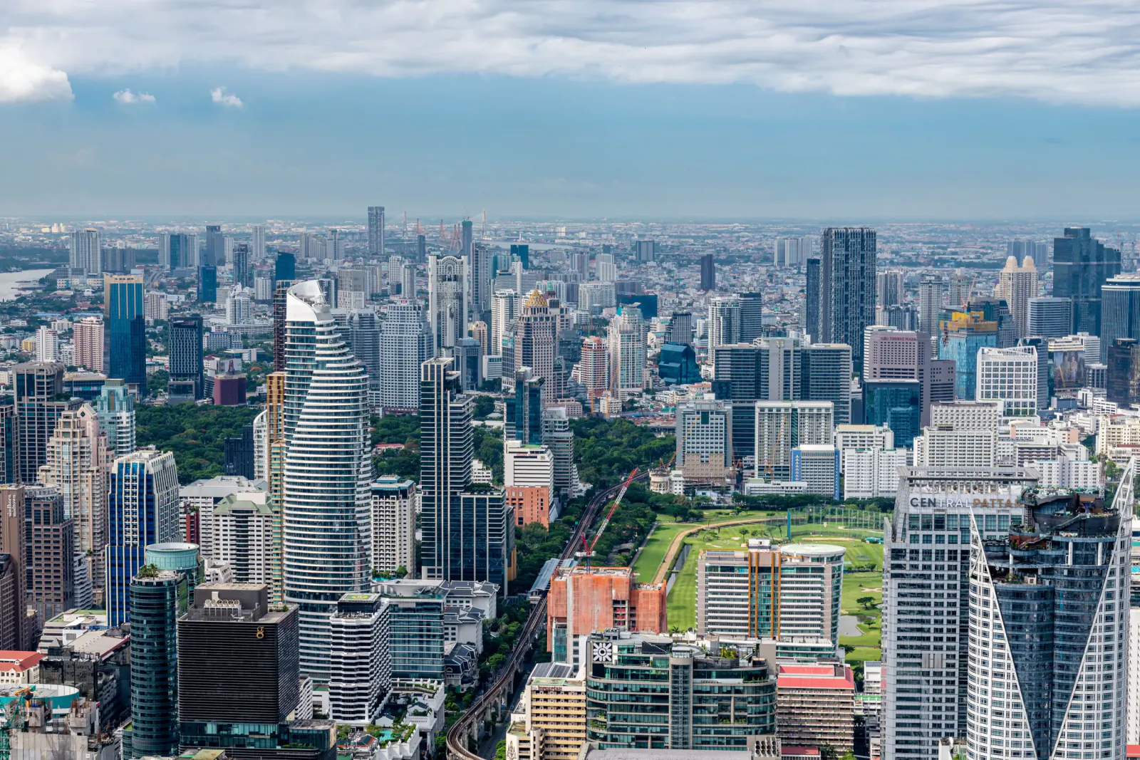 Aerial view of Bangkok city skyline with modern skyscrapers, green parks, and a train line under a partly cloudy sky