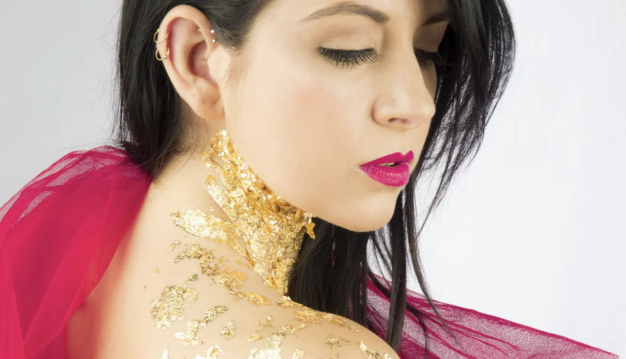 Woman with closed eyes wearing gold leaf jewelry and magenta lipstick, draped in sheer red fabric