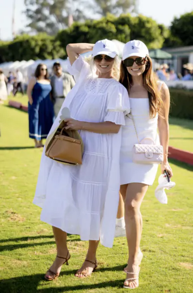 Two women in white outfits and caps pose on a sunny grass field at a polo event, one in a tiered dress holding a brown handbag and the other in a short dress with a white quilted purse.