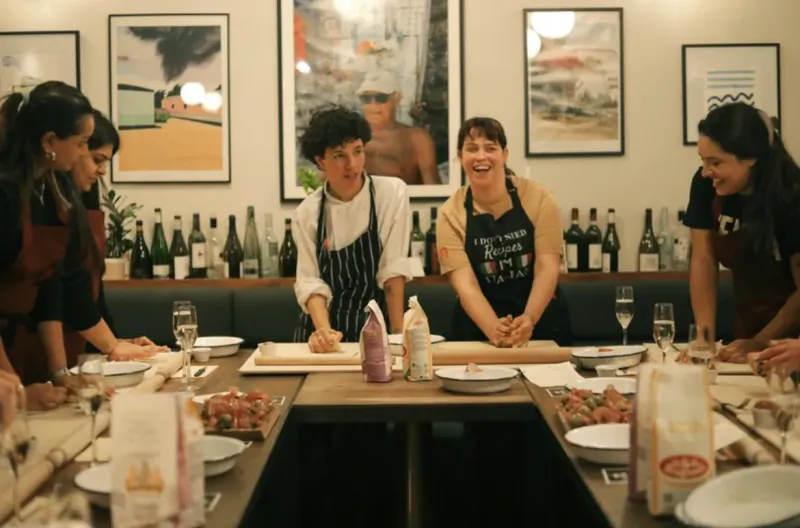 Five women in aprons gather around a long table in a restaurant, smiling and preparing pasta dough together