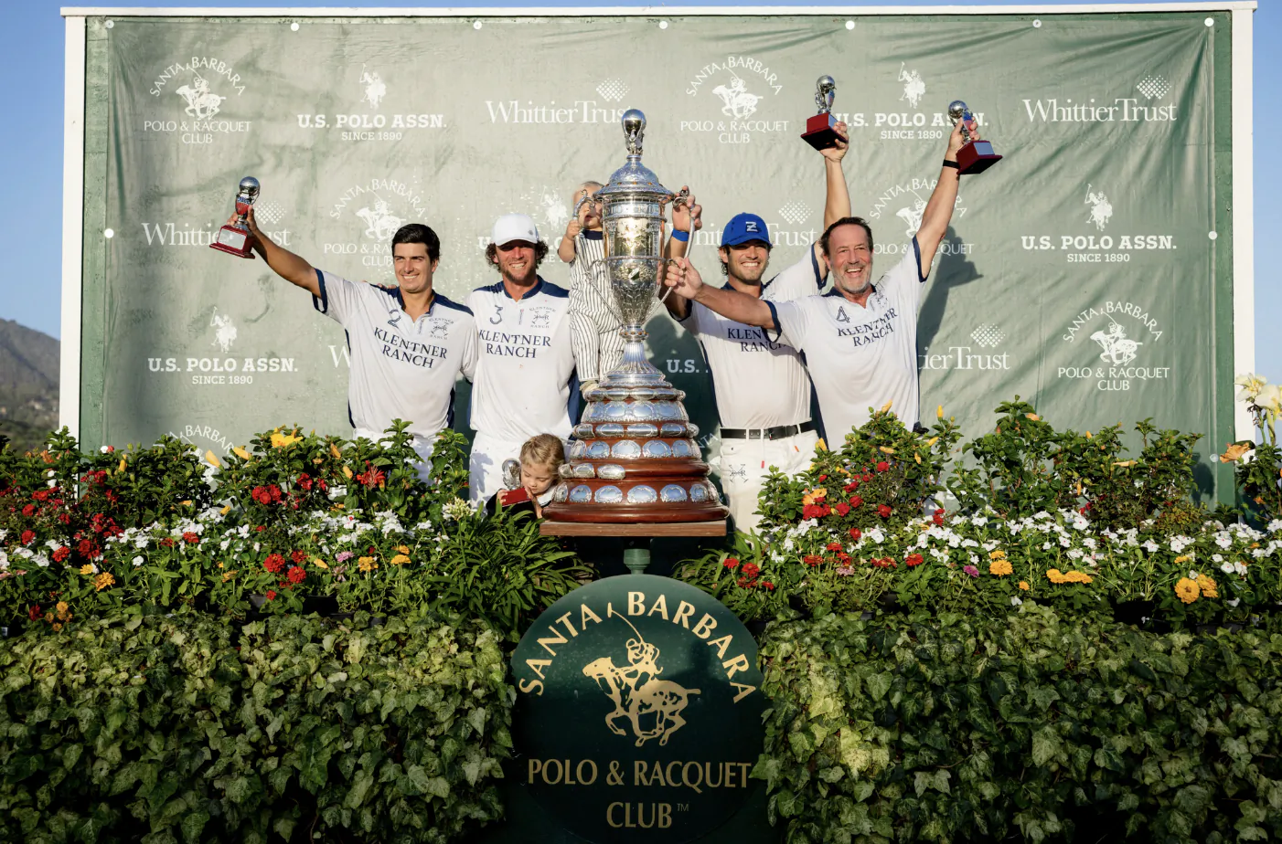 Four polo players in white jerseys celebrate with the Pacific Coast Open trophy, raising polo mallets in front of a U.S. Polo Assn. banner at Santa Barbara Polo & Racquet Club