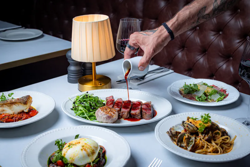 Tattooed hand pouring red wine over sliced steak at a restaurant table with multiple gourmet dishes