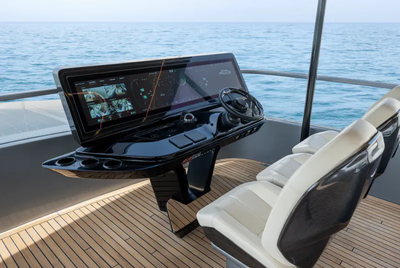 Modern yacht helm station with large touchscreen displays, steering wheel, and white leather seats overlooking calm blue ocean