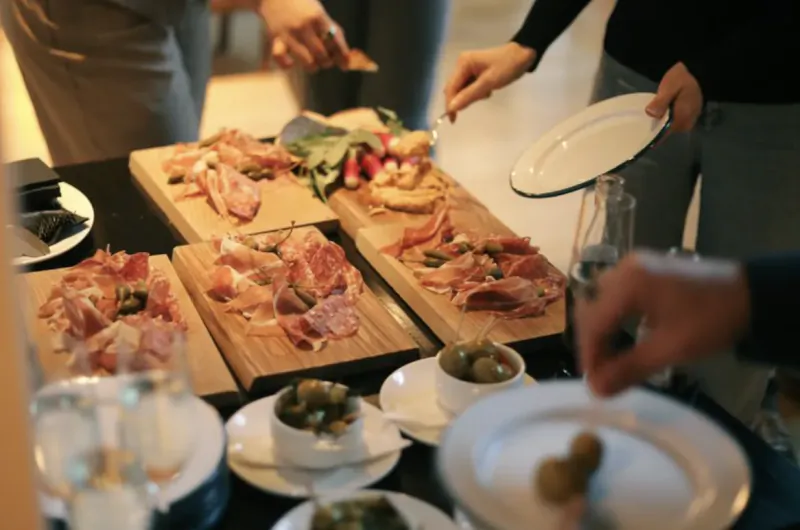 People serving themselves cured meats, olives, and appetizers from wooden boards at a buffet table