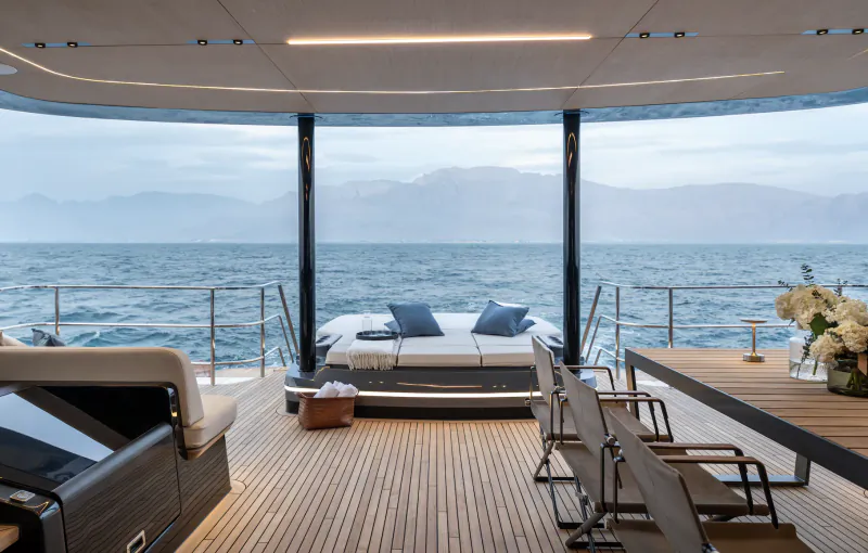 Luxury yacht deck with large bed facing panoramic ocean and mountain view, dining table and chairs on wooden flooring