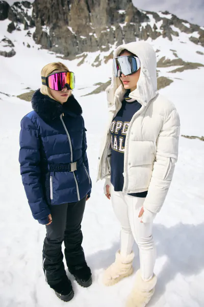 Two women in colorful ski goggles and puffer jackets stand on a snowy mountain slope