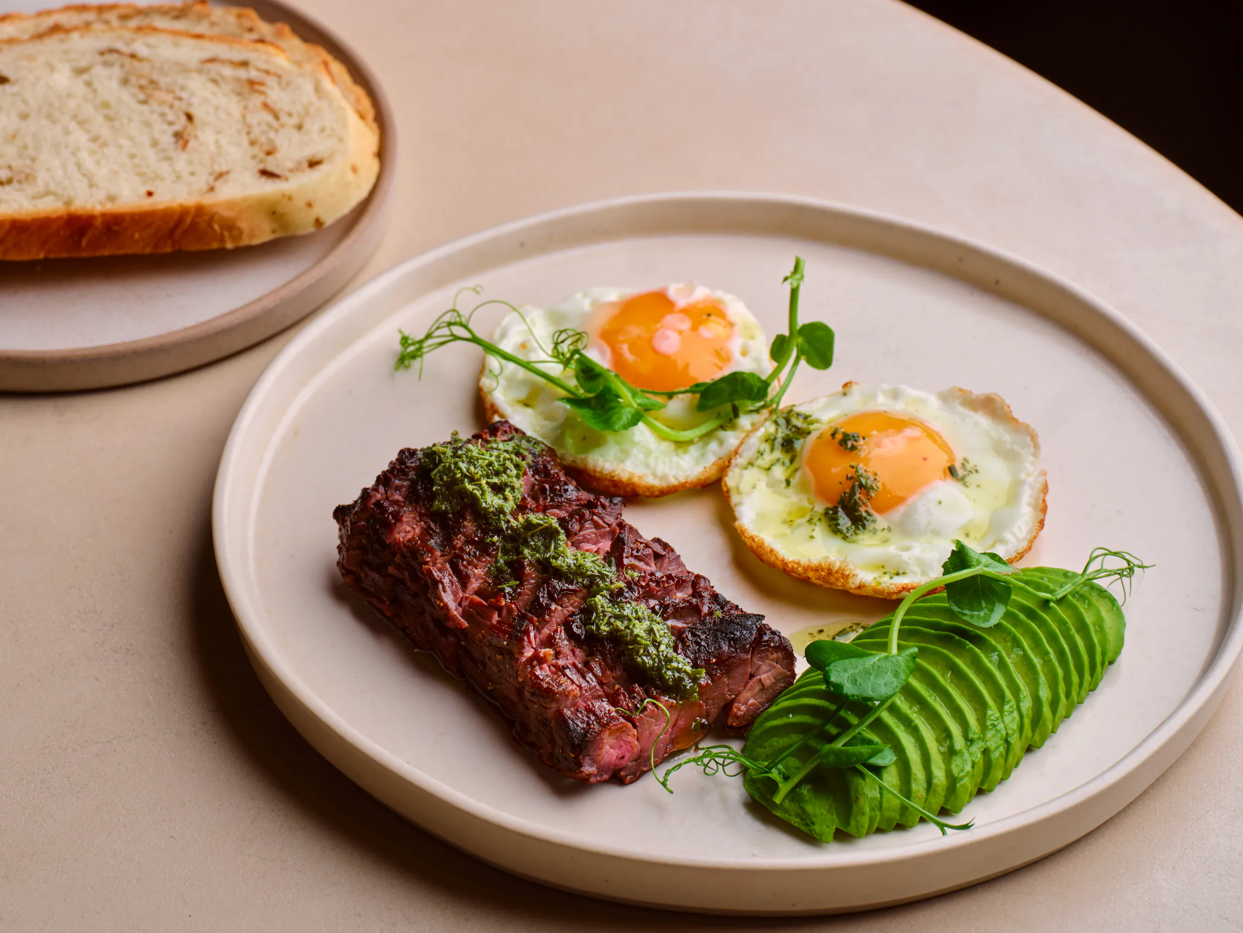 Plate with seared steak topped with green sauce, two sunny-side-up eggs, sliced avocado, and microgreens, with toast on the side