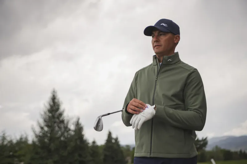 Man in green jacket and blue cap holding golf club on overcast course