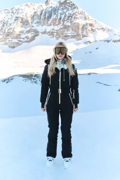 Woman in black ski suit with white trim and goggles standing on snowy mountain slope