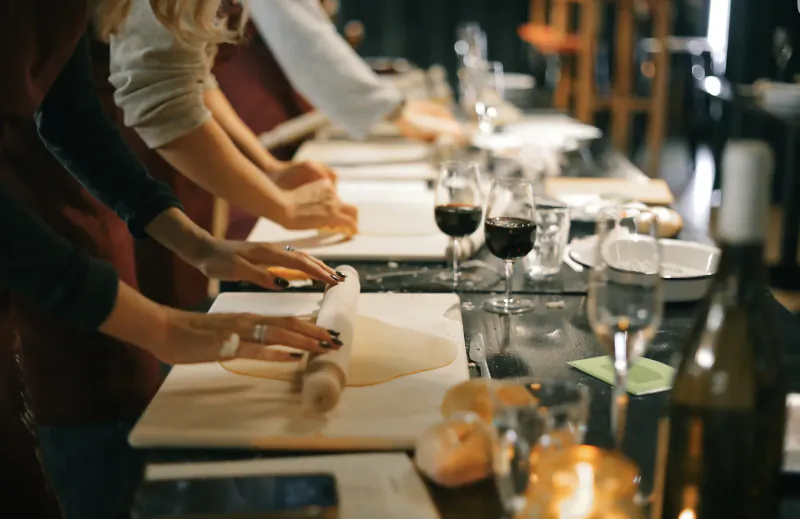 People rolling fresh pasta dough at a long table with wine glasses and bread during a pasta-making class