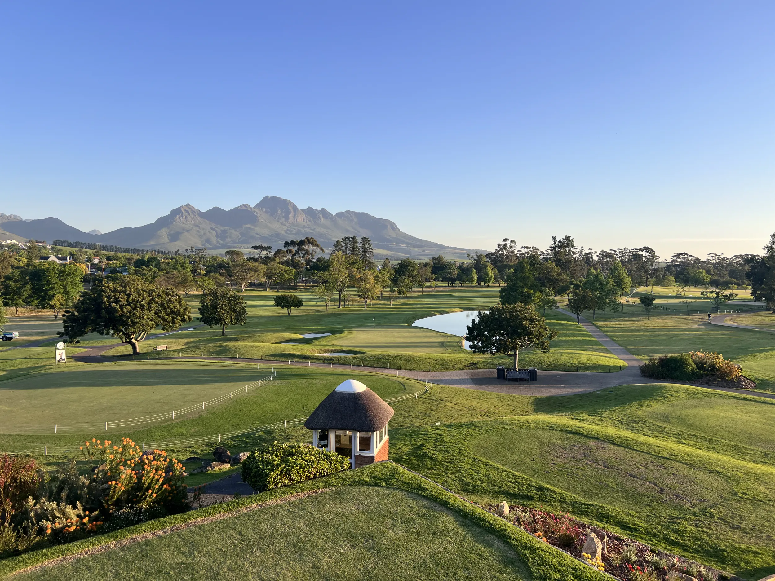 Scenic golf course with thatched hut, pond, and mountain backdrop under clear blue sky