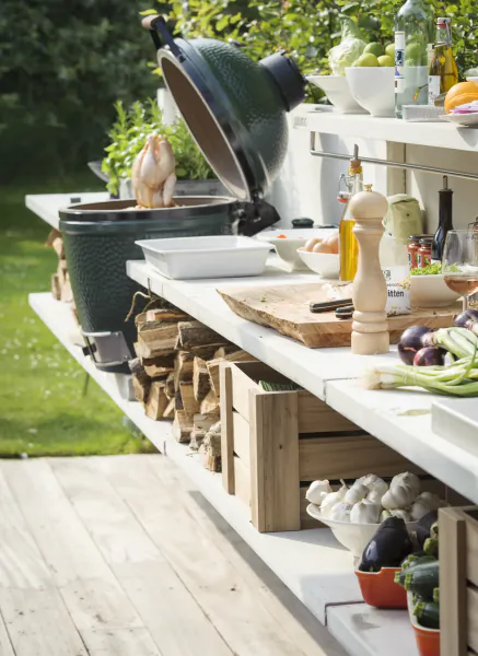 Outdoor kitchen with open green ceramic grill, raw chicken, fresh vegetables, cutting board and wine on white counter