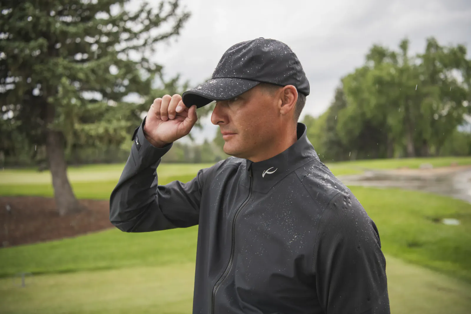 Man in black rain jacket and cap adjusts brim while standing on rainy golf course