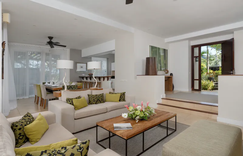 Bright open-plan living room with beige sofas, green cushions, wooden coffee table and modern kitchen in a luxury villa