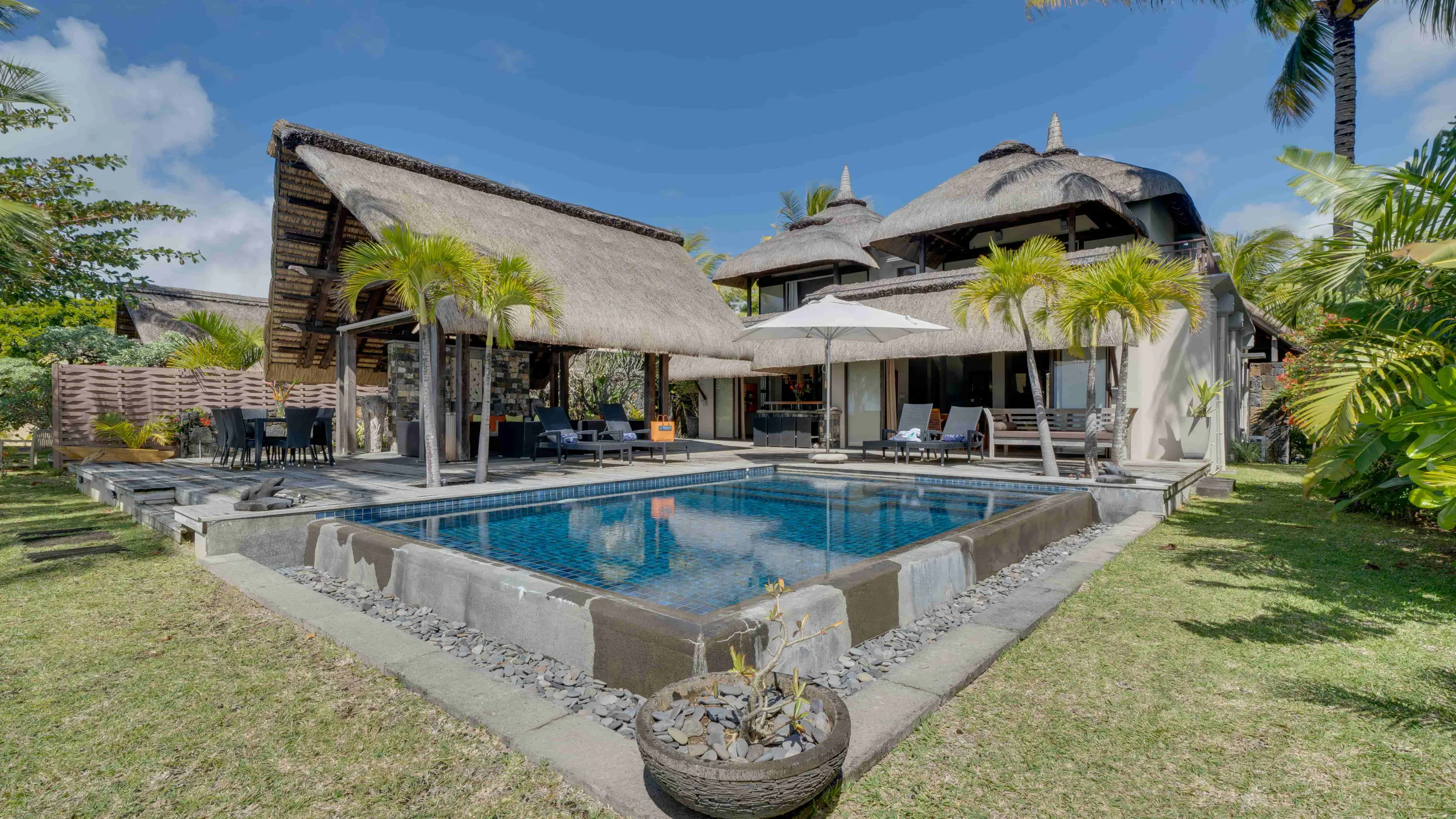 Luxury villa with thatched roofs, swimming pool, patio furniture and tropical garden under blue sky