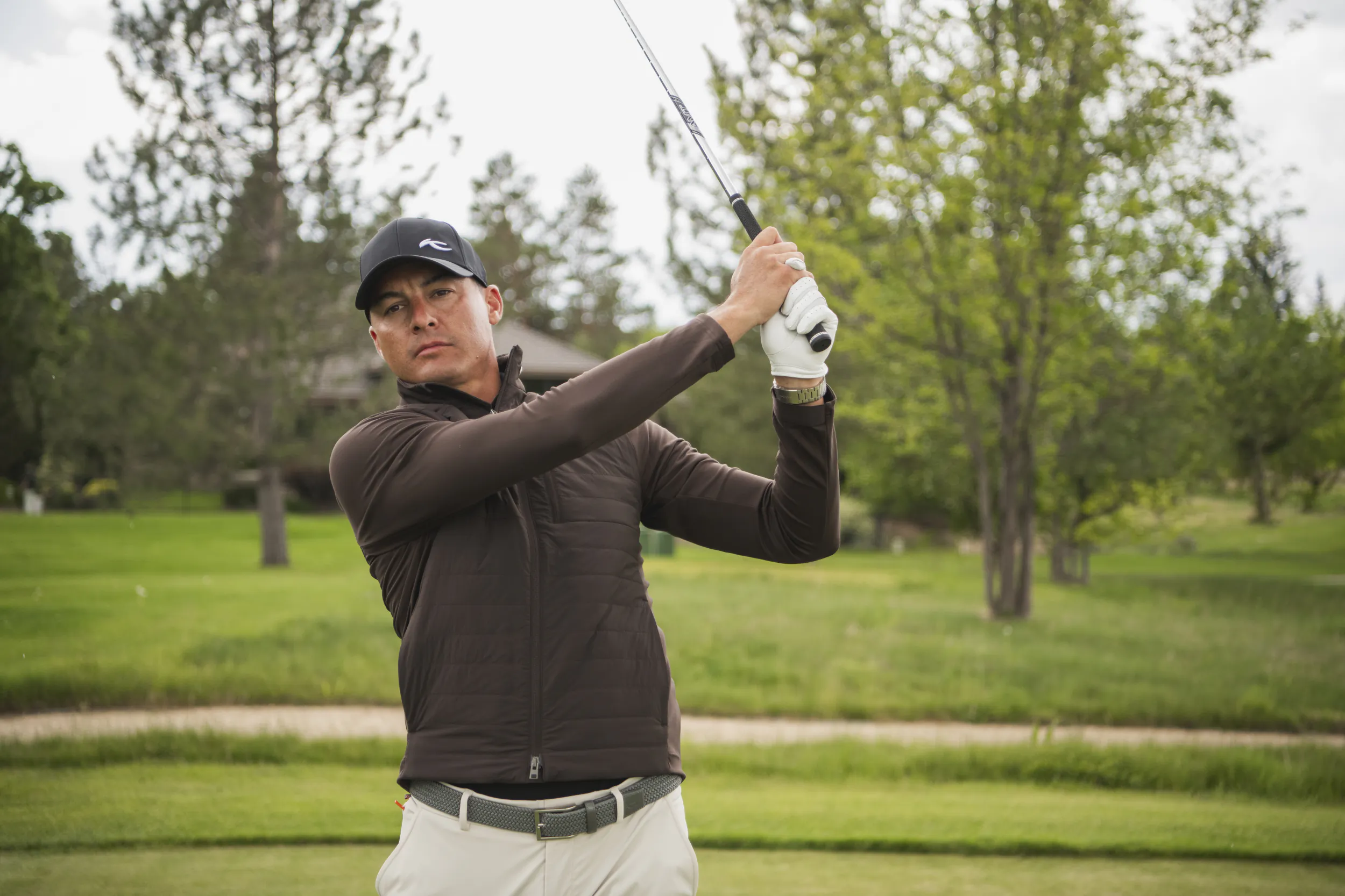 Male golfer in brown jacket and cap follows through on a swing on a lush green course
