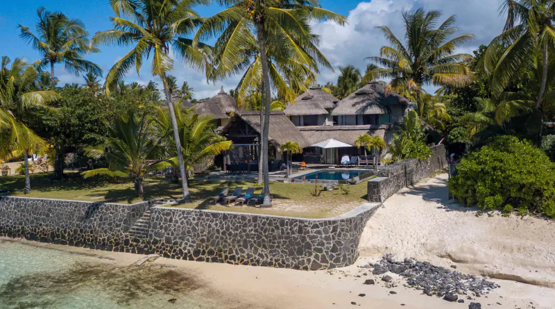 Luxury beachfront villa with thatched roofs, palm trees, pool, and stone wall overlooking sandy beach in Mauritius