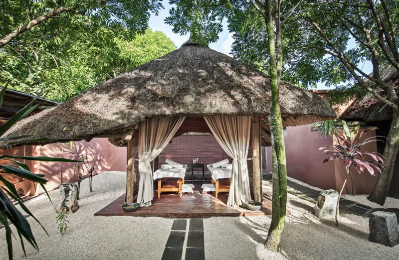 Thatched-roof African-style lounge with two white armchairs on a red rug, set in a sandy garden with pink walls and lush trees