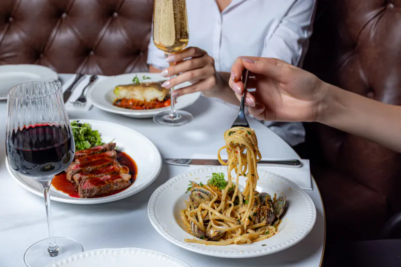 Woman in white shirt twirling seafood pasta with fork at upscale restaurant table with steak, wine, and champagne
