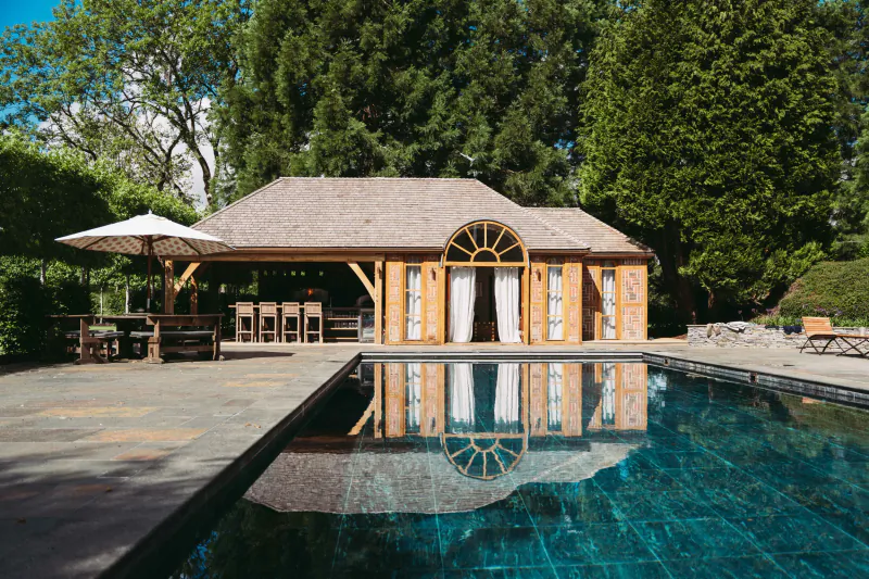 Wooden poolside building with arched window and white curtains reflected in turquoise water, surrounded by trees and outdoor seating