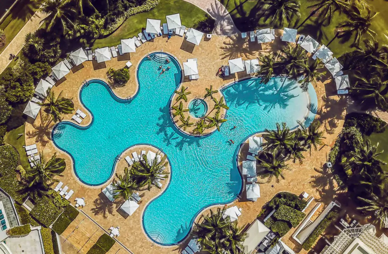 Aerial view of a large, winding resort pool with palm trees, lounge chairs, and cabanas at Continuum Luxury Residences