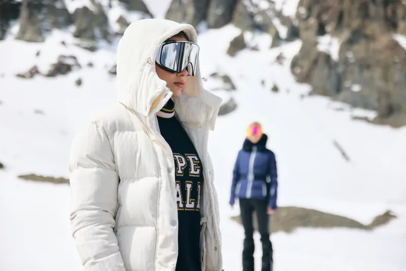 Woman in white puffer jacket and ski goggles standing in snowy mountain landscape with another skier in background