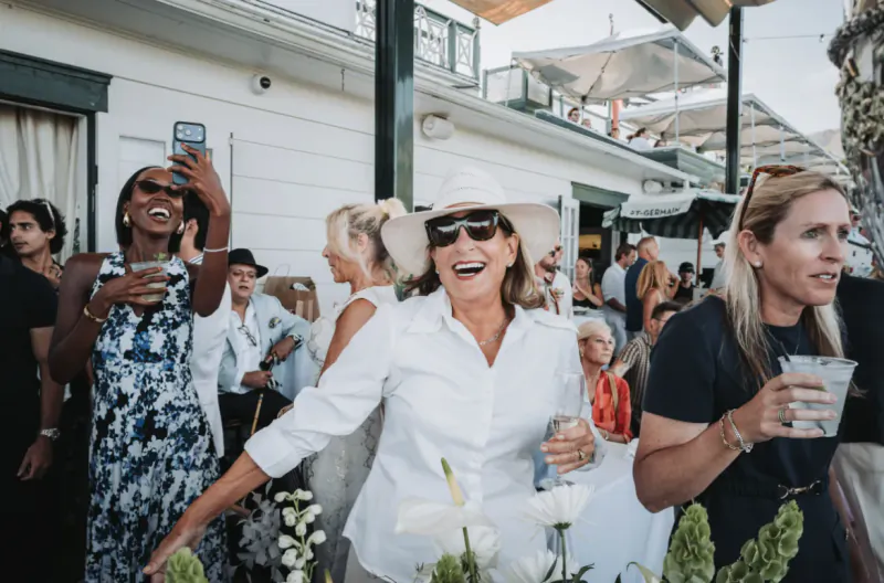 Woman in white hat and shirt laughing at outdoor party, holding wine glass, surrounded by guests with drinks
