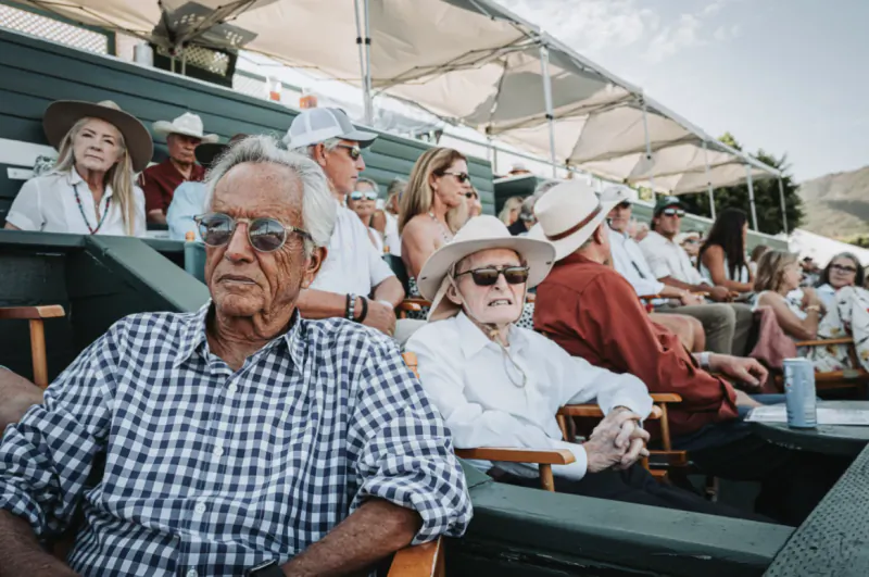 Elderly couple in sunglasses seated in outdoor stadium seating, man in checkered shirt and woman in white hat, with spectators behind